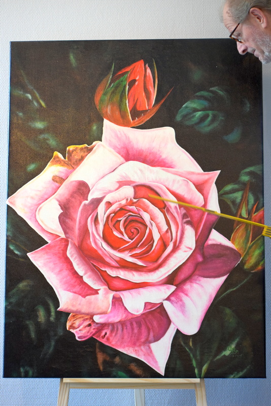 Gemälde einer blühenden Rose mit einer Knospe auf einer Staffelei. Am oberen Bildrand schaut das Gesicht eines bärtigen Mannes hervor.