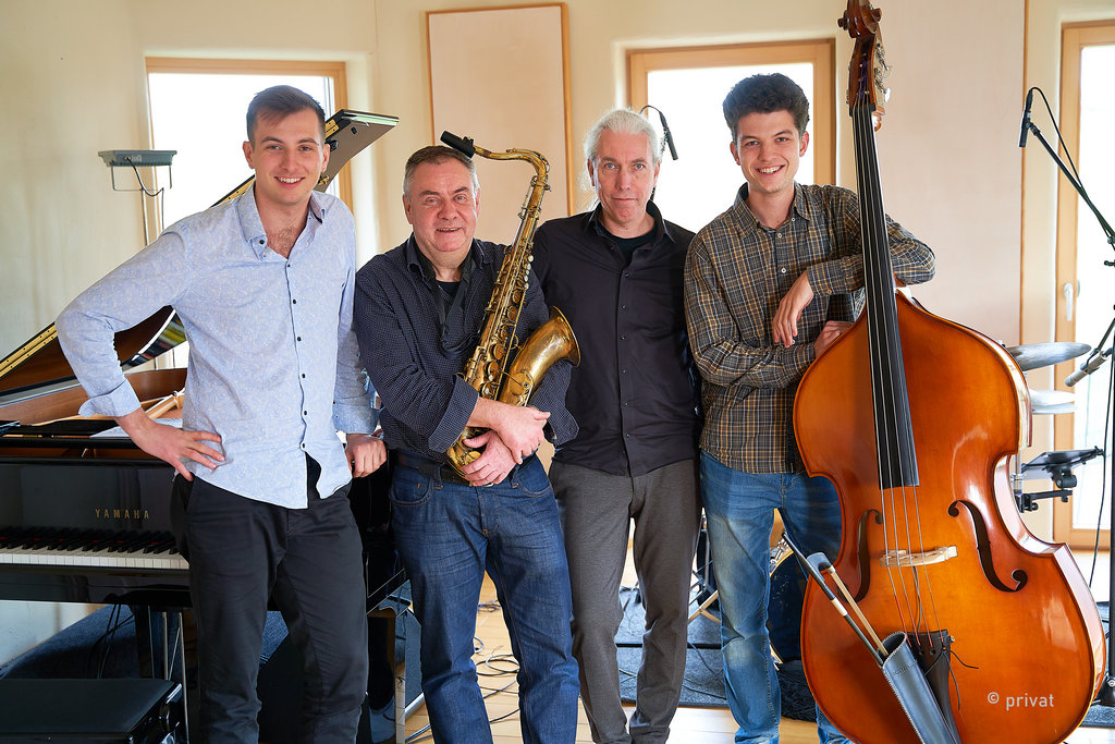 Vier Musiker mit Saxophon und Bass neben einem Klavier