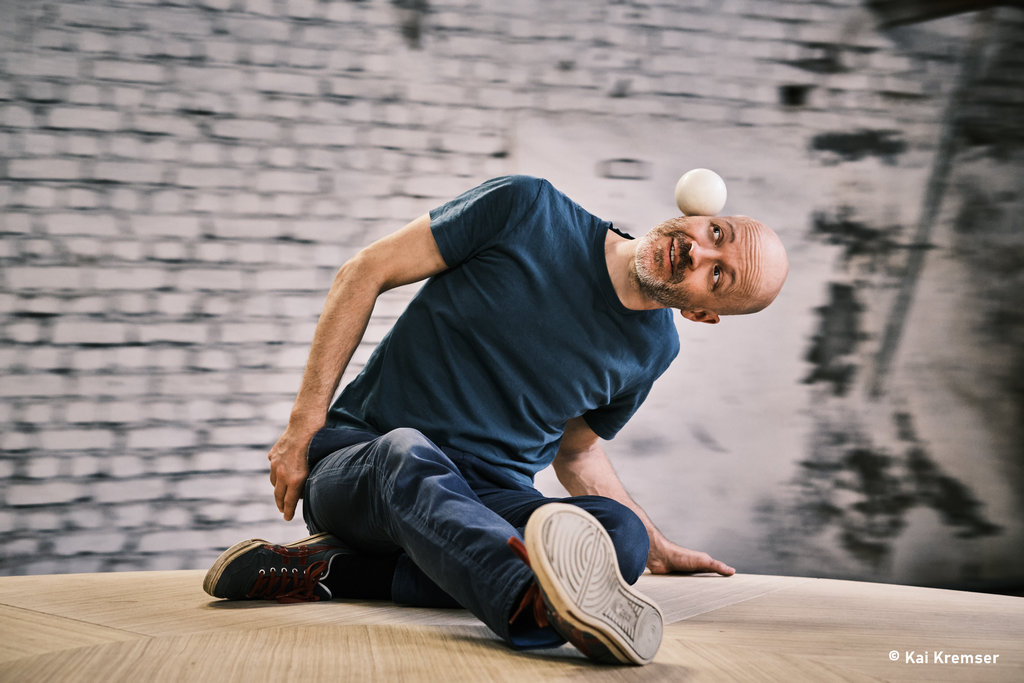 Ein Mann jongliert sitzend einen kleinen weißen Ball auf seinem Kopf